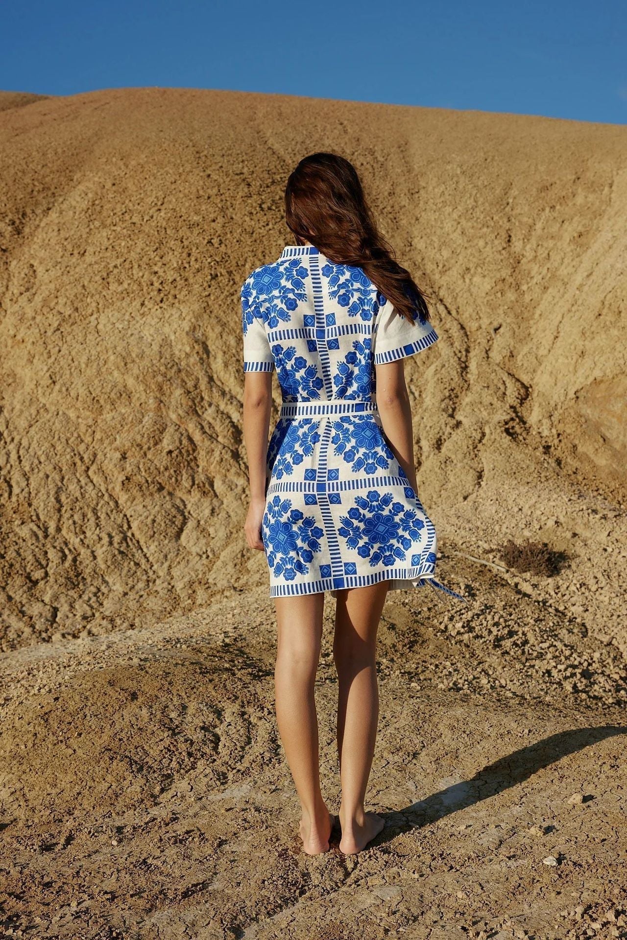 A woman stands with her back to the camera in a desert landscape, wearing a white and blue floral print shirt dress.