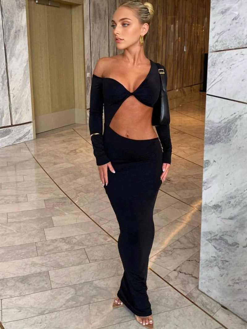 A woman in a black one-shoulder, cut-out maxi dress with a twist-front detail stands in a hallway with marble walls.