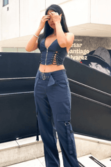 A young woman stands on stairs outdoors, adjusting her hair and posing for a photo in a navy blue lace-up crop top and cargo pants.
