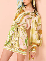 woman leaning against a pink wall wearing a white, green, orange, blue, pink, and yellow patterned silk shirt dress with a tie waist and puffed sleeves