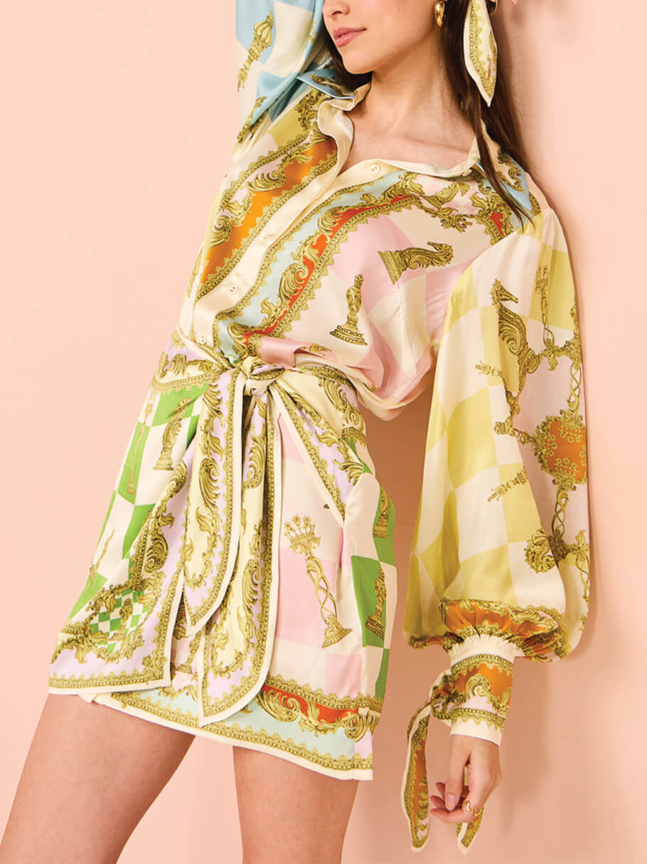woman leaning against a pink wall wearing a white, green, orange, blue, pink, and yellow patterned silk shirt dress with a tie waist and puffed sleeves