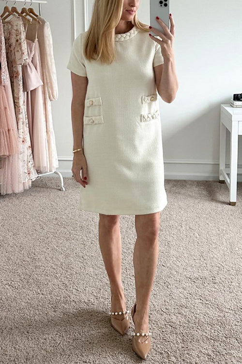 A woman taking a mirror selfie in a cream-colored tweed shift dress with short sleeves and decorative pockets, wearing pearl-embellished heels.