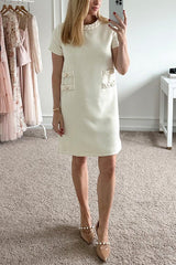 A woman taking a mirror selfie in a cream-colored tweed shift dress with short sleeves and decorative pockets, wearing pearl-embellished heels.