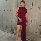 A woman in a burgundy draped dress with an asymmetrical hemline and a thigh-high slit, posing against a concrete wall.