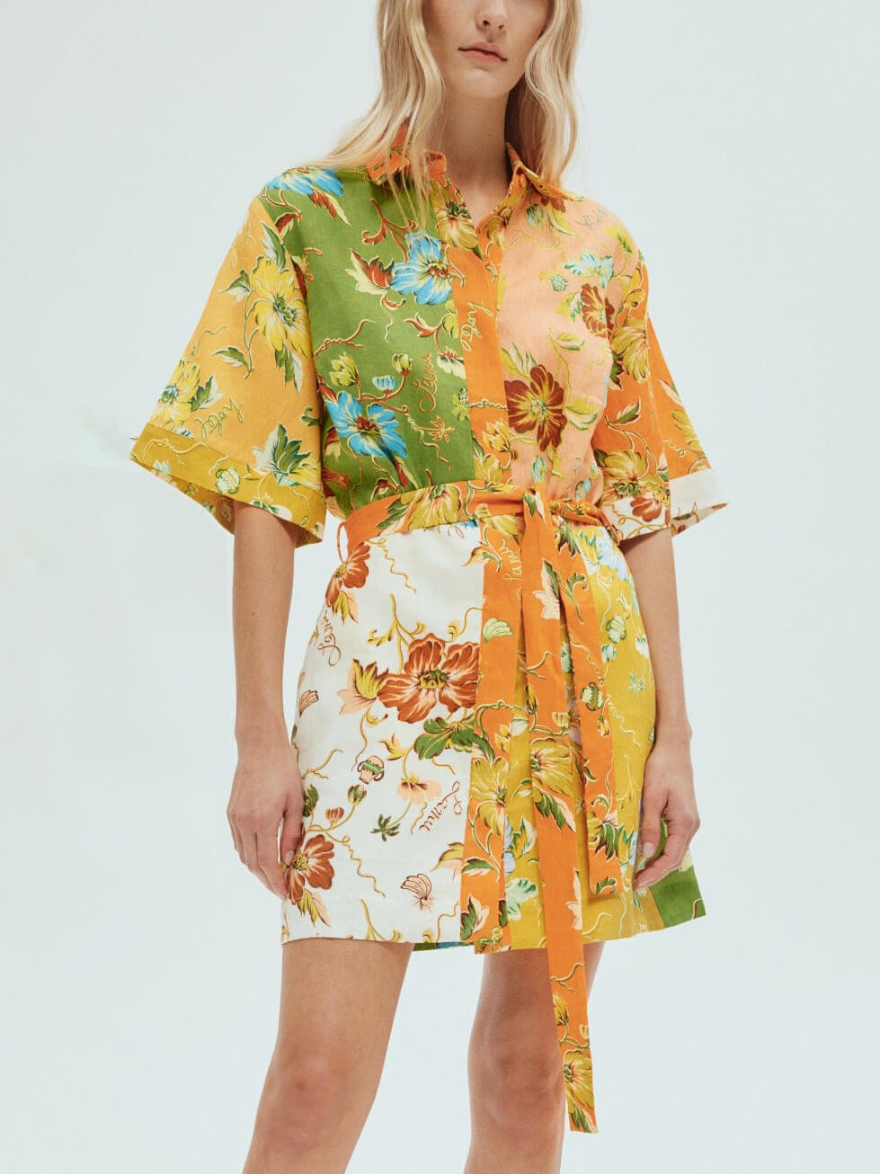 A woman wearing a short-sleeved, collared shirt dress with a floral print and a belted waist, standing against a white background.
