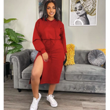 A woman in a red hooded midi dress with a high slit, white sneakers, and hoop earrings, posing indoors in front of a gray couch.