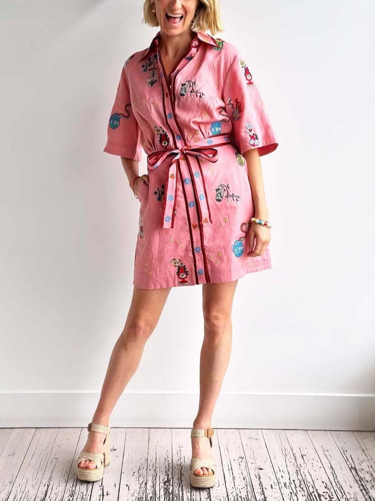 A woman wearing a pink shirt dress with embroidered designs and a belt, standing on a wooden floor.