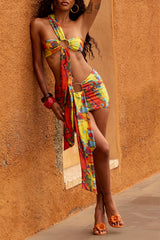 Woman leaning against an orange wall wearing a colorful abstract print bikini top and mini skirt with a draped scarf detail.
