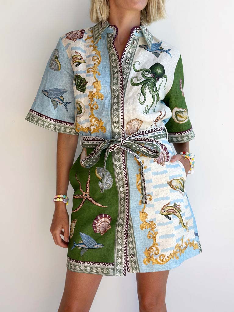 Close-up of a woman wearing a collared shirt dress with a detailed oceanic print. The dress has short sleeves and a belt tied at the waist.