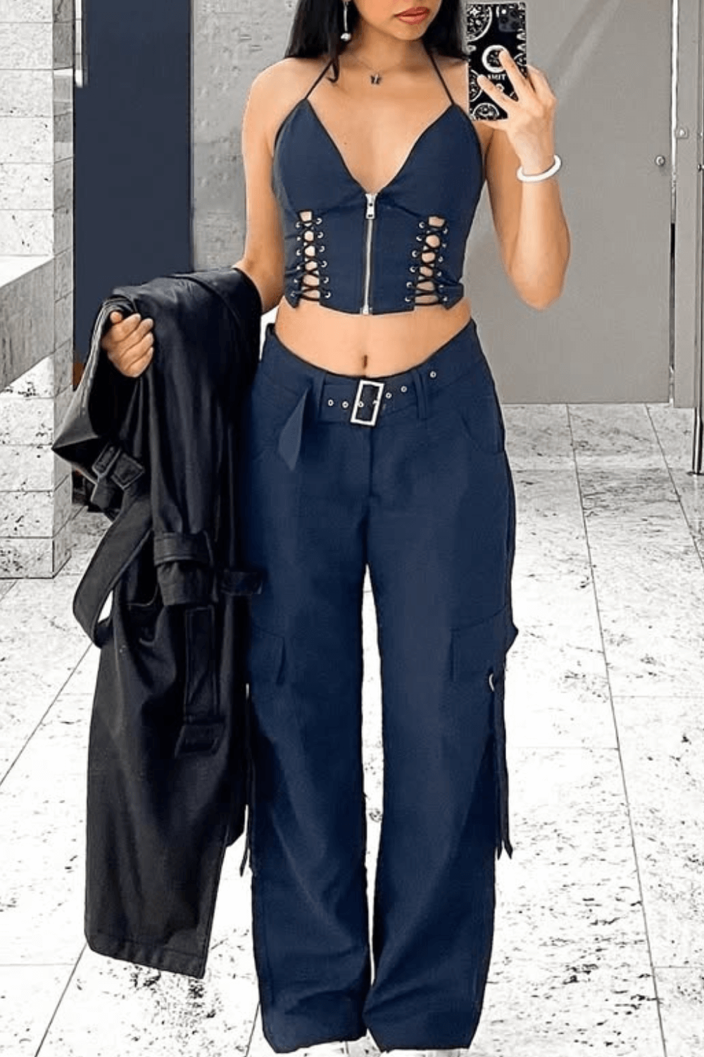 A young woman stands in an indoor space, holding a black jacket over her arm and posing for a photo with her phone.