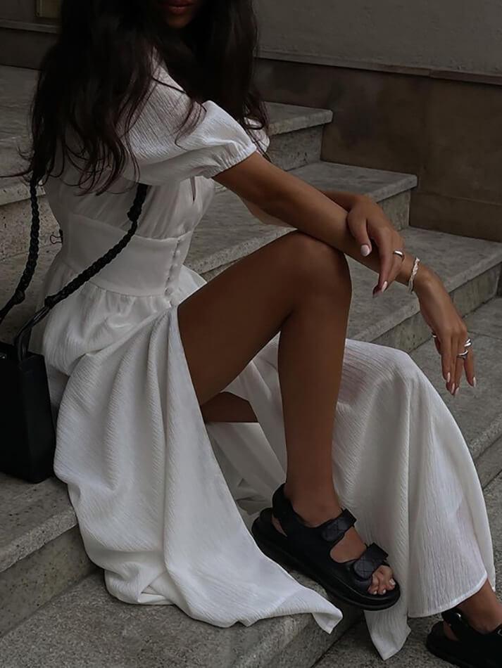 Close-up of a woman in a white maxi dress sitting on steps, showing the slit and sandals.