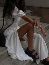 Close-up of a woman in a white maxi dress sitting on steps, showing the slit and sandals.