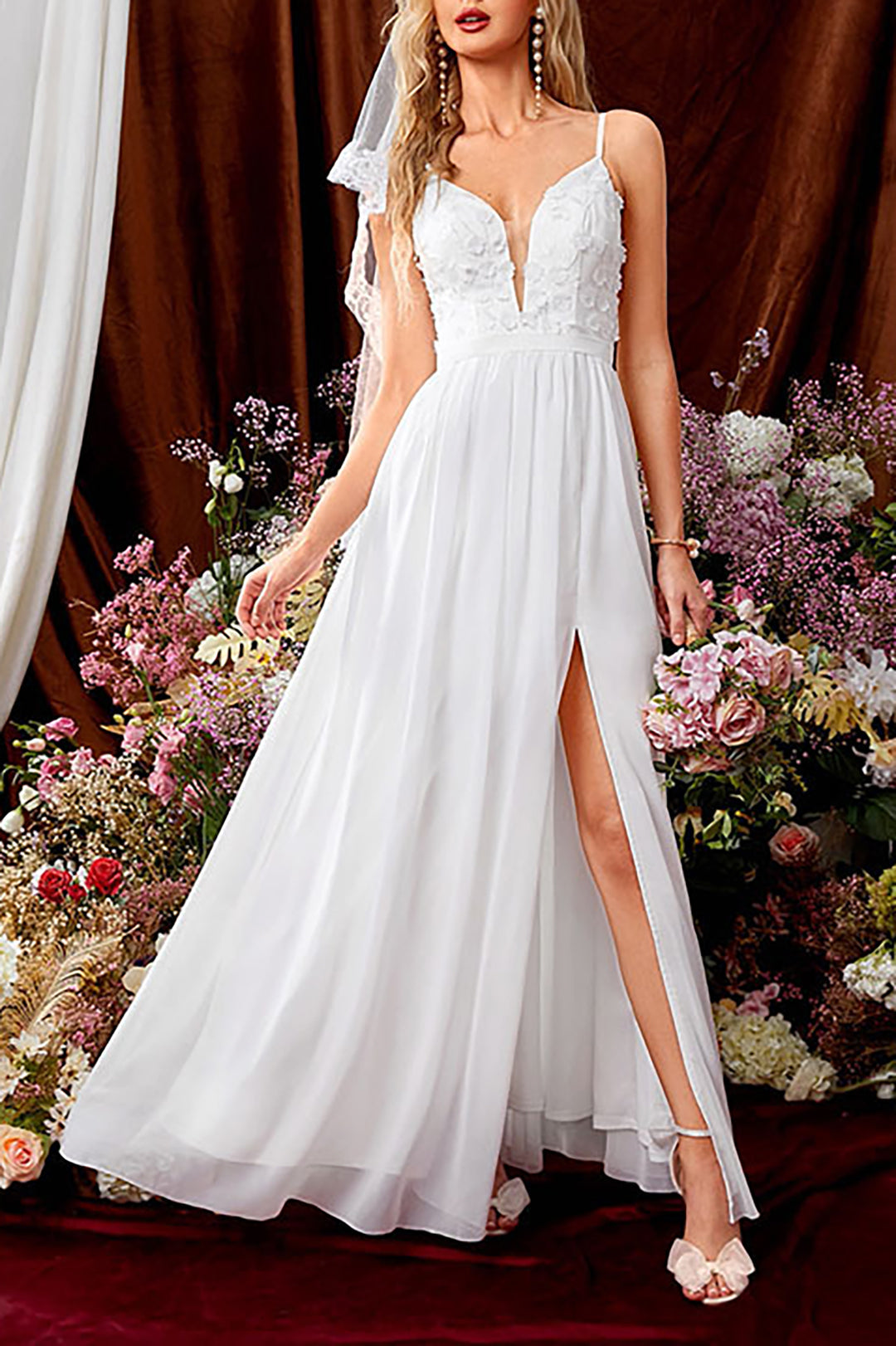 Full-body shot of a woman in a white wedding dress with a high slit and floral appliques, standing in front of a floral backdrop.