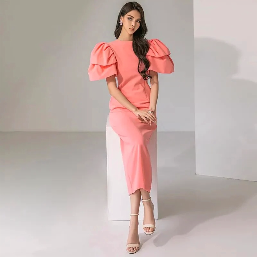 woman in a coral pink midi dress with dramatic puff sleeves sitting on a white cube