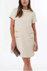 A woman wearing a cream-colored tweed shift dress with short sleeves and decorative pockets, holding a tortoiseshell clutch.