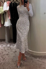 A woman wearing a long-sleeved, silver sequined midi dress and clear embellished heels, standing in a dressing room.