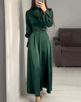 Woman wearing a long emerald green satin dress with a high neckline, cinched waist, and ruffled cuffs, standing in a room with a white wall and a brown sofa.