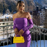 A woman in a purple off-the-shoulder ruched dress stands on a balcony with a snowy mountain town in the background.