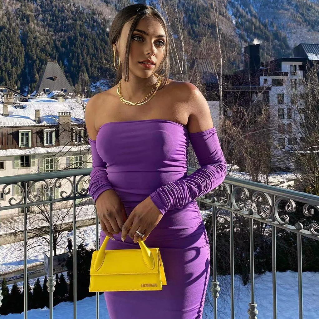 A woman in a purple off-the-shoulder ruched dress stands on a balcony with a snowy mountain town in the background.