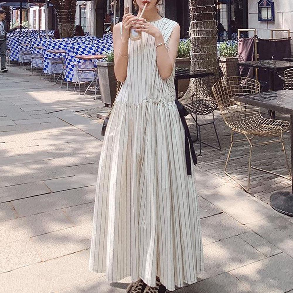 Woman wearing a long white sleeveless dress with black vertical stripes, holding a drink, standing on a street with outdoor seating.