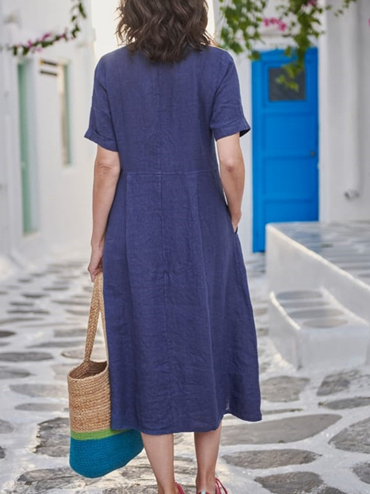 Back view of a woman wearing a navy blue linen midi dress with pockets. She is holding a straw tote bag with a teal and green stripe.