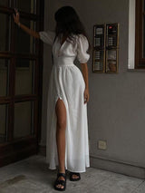 A woman in a white maxi dress with a high slit and black sandals stands by a door.