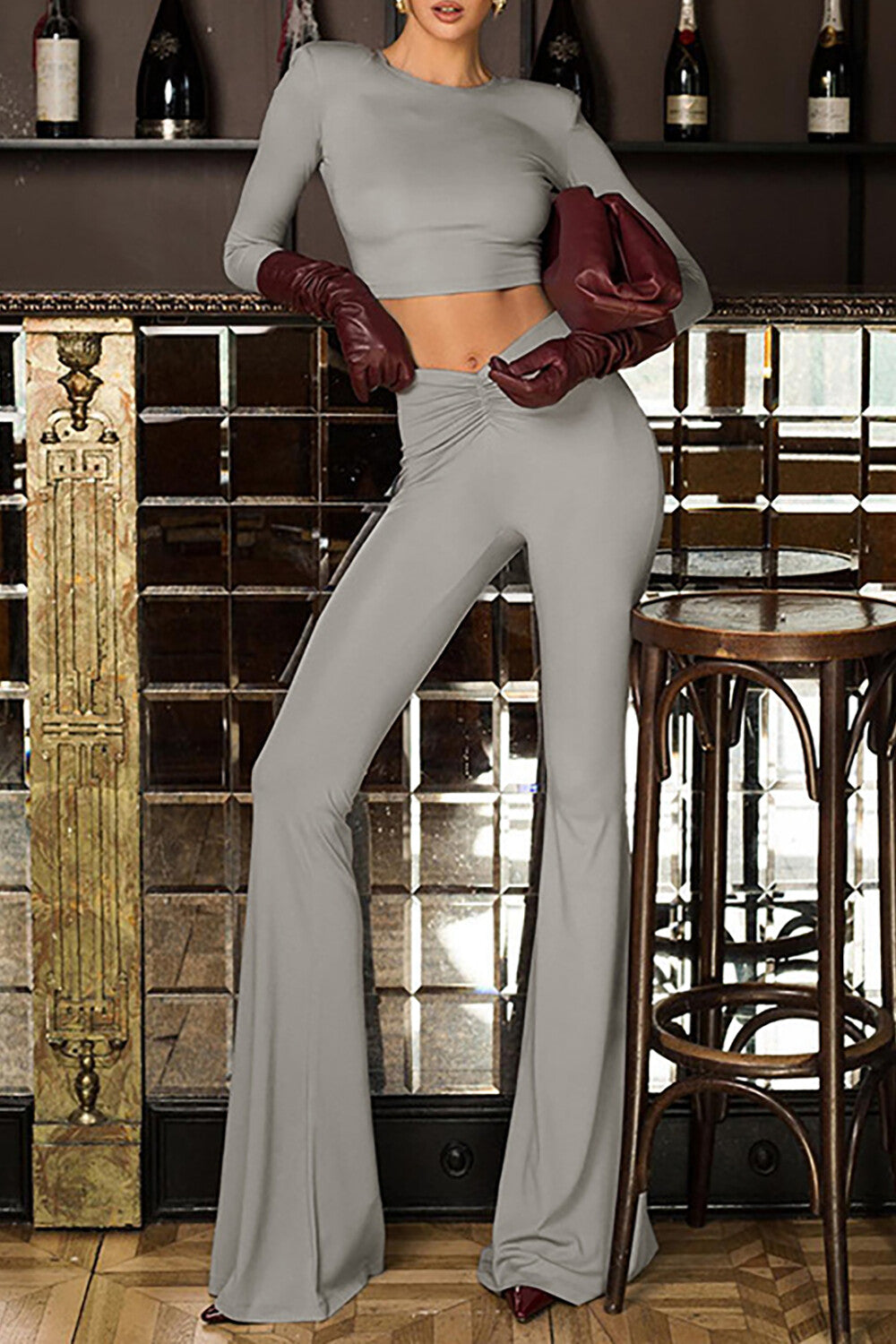 Woman wearing a gray cropped long-sleeve top and gray flare pants, accessorized with burgundy gloves and a burgundy clutch bag, posing in a bar setting.