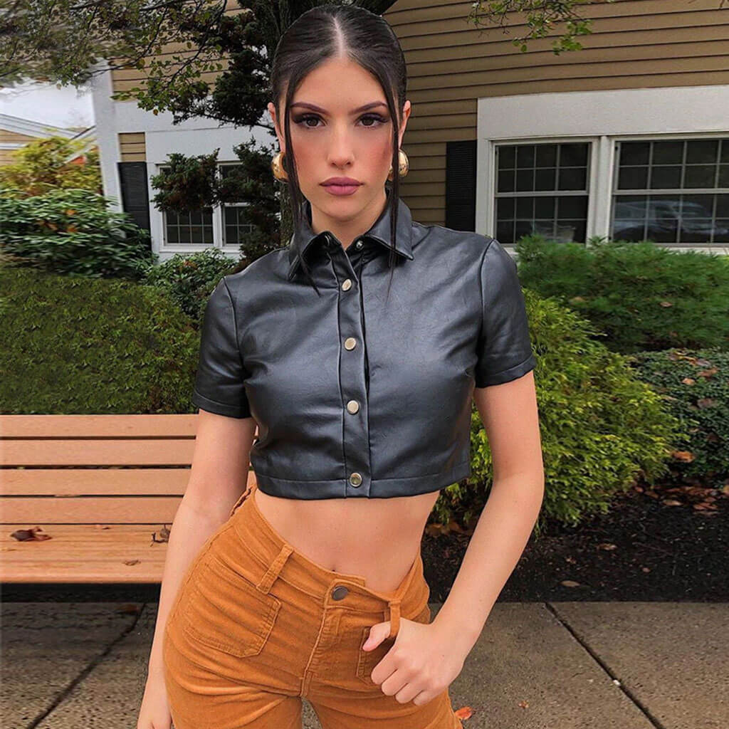 A young woman with dark hair pulled back stands outside, wearing a black faux leather cropped shirt with gold buttons and orange corduroy pants.