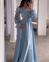 A woman in a light blue satin dress with dramatic puff sleeves and a square neckline is taking a mirror selfie.