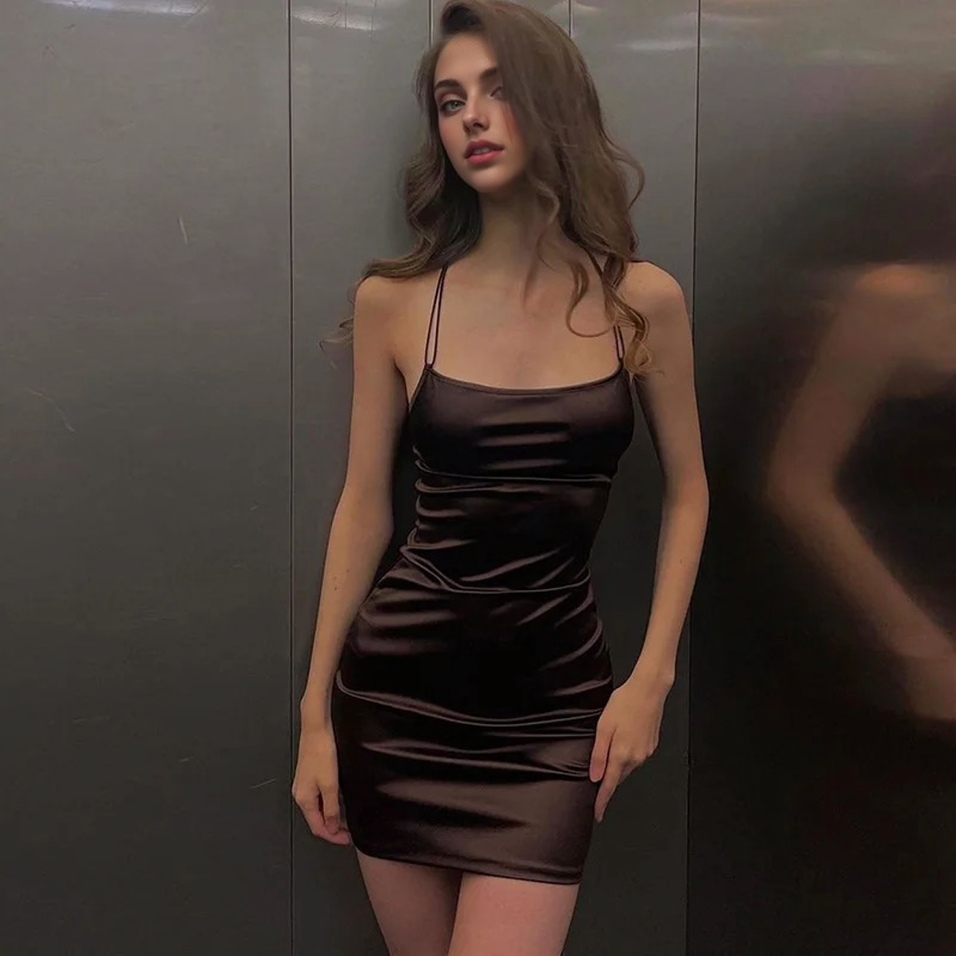 A woman in a brown satin mini dress leans against a metallic wall.