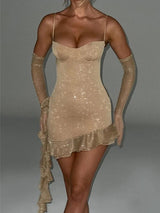 A woman wearing a short, champagne-colored, sequined dress with spaghetti straps, a sweetheart neckline, ruffle trim, and an asymmetrical hem. She is also wearing long, sheer champagne-colored gloves.