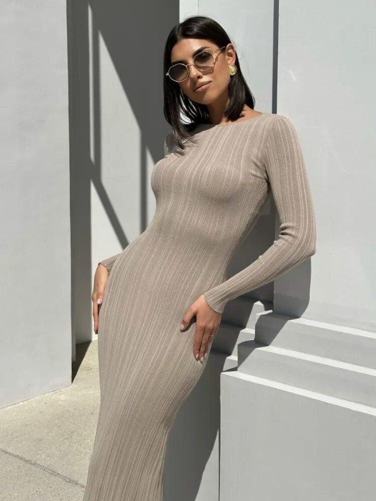 A woman with dark bob haircut and sunglasses leans against a white wall in a beige ribbed knit maxi dress.