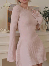 A woman wearing a light pink knit mini dress with long bell sleeves and a scoop neckline, posing indoors.