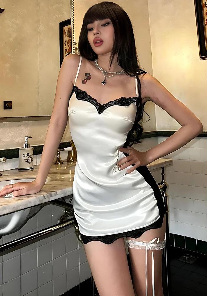 Woman in a white satin slip dress with black lace trim, leaning on a marble countertop.