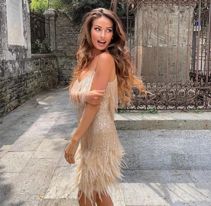 Woman in a gold sequined and feathered mini dress with spaghetti straps, posing with her arm on her hip outdoors.