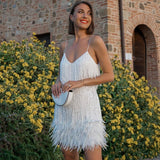 Woman in a white sequined and feathered mini dress with spaghetti straps, standing outdoors in front of a brick building and yellow flowers.