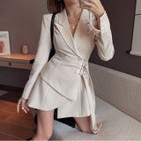 A woman poses in a beige blazer dress with a wrap skirt and belted waist.