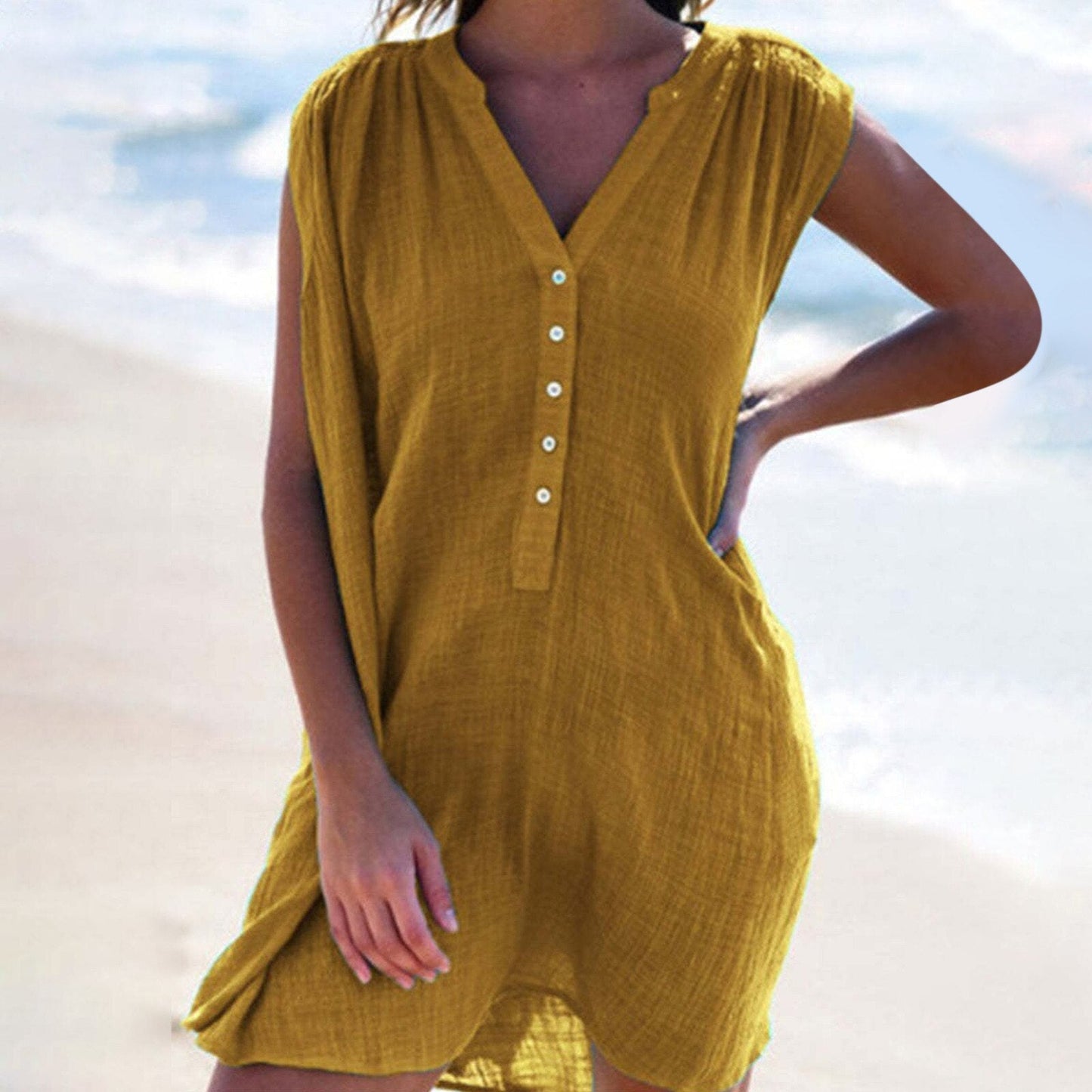 A woman wearing a mustard yellow sleeveless dress with a v-neck and button-down front, standing on a beach.
