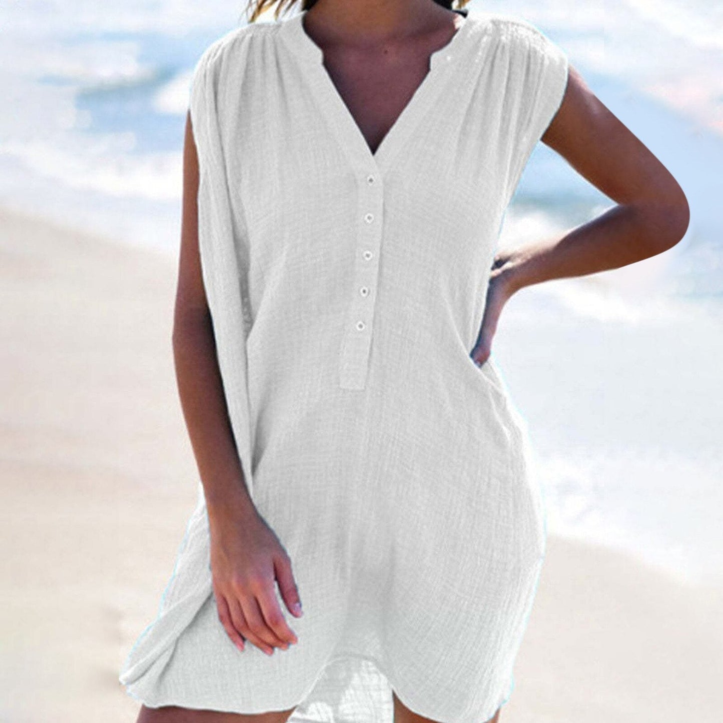 A woman wearing a white sleeveless dress with a v-neck and button-down front, standing on a beach.