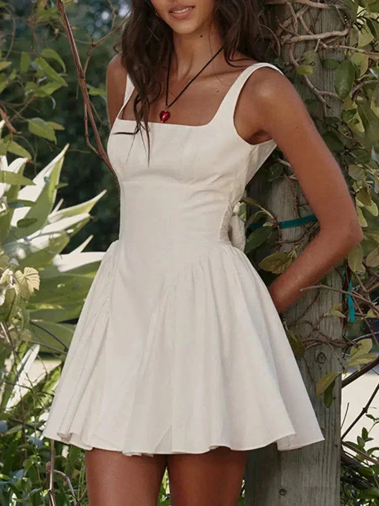 woman wearing a white sleeveless dress with a square neckline and a bow detail on the back, standing in a garden