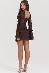 Back view of a woman wearing a short, brown, long-sleeved ruched dress with a square neckline and bell sleeves.