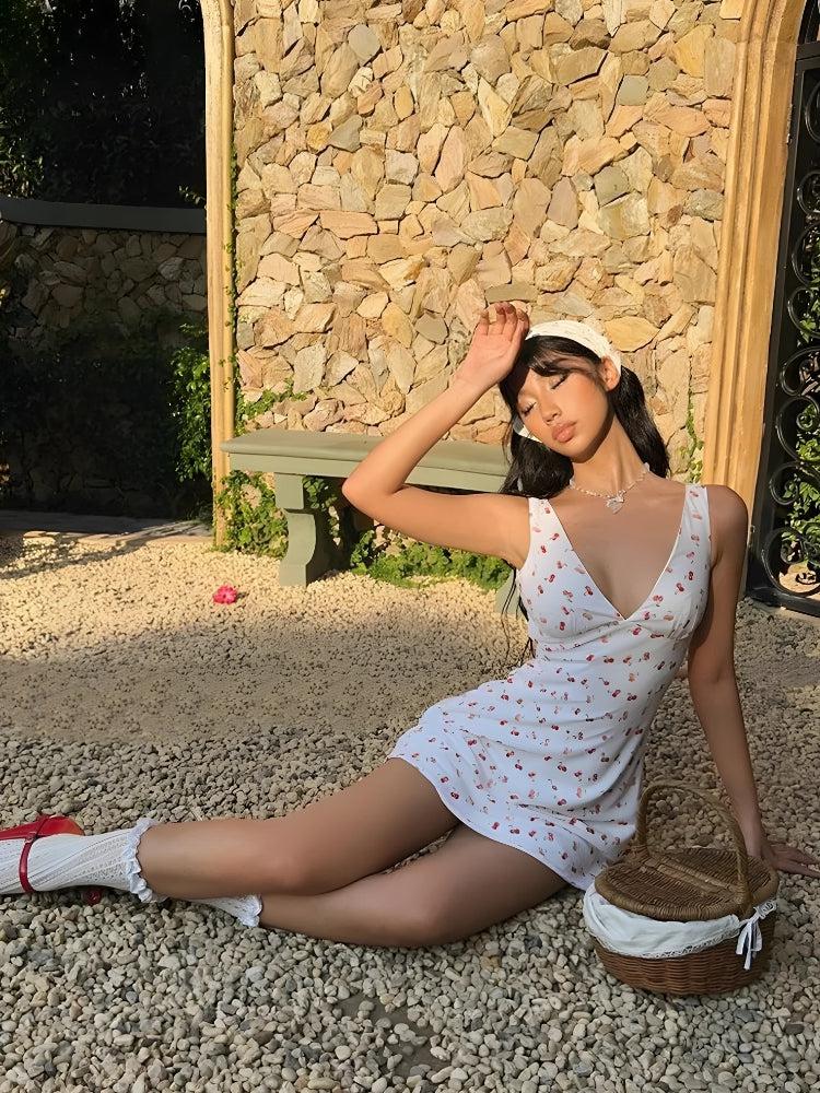 A woman in a white cherry-print mini dress, white bandana, red mary jane heels, and white ruffled socks, sitting on the ground