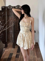 A woman wearing a cream corset-style mini dress with spaghetti straps and a ruffled skirt, posing with her hand on her head in a room with wooden floors and a decorative screen.