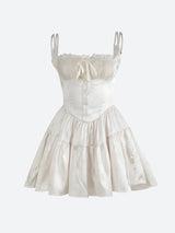 A white corset-style mini dress with spaghetti straps, lace trim, a tie front, button detailing, and a ruffled tiered skirt, shown against a white background.