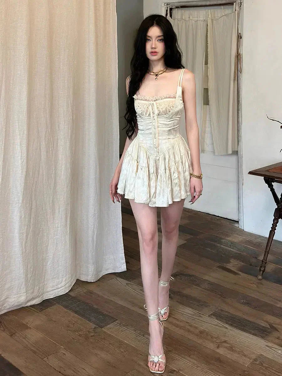 A woman wearing a cream corset-style mini dress with spaghetti straps and a ruffled skirt, standing in a room with wooden floors and sheer curtains.