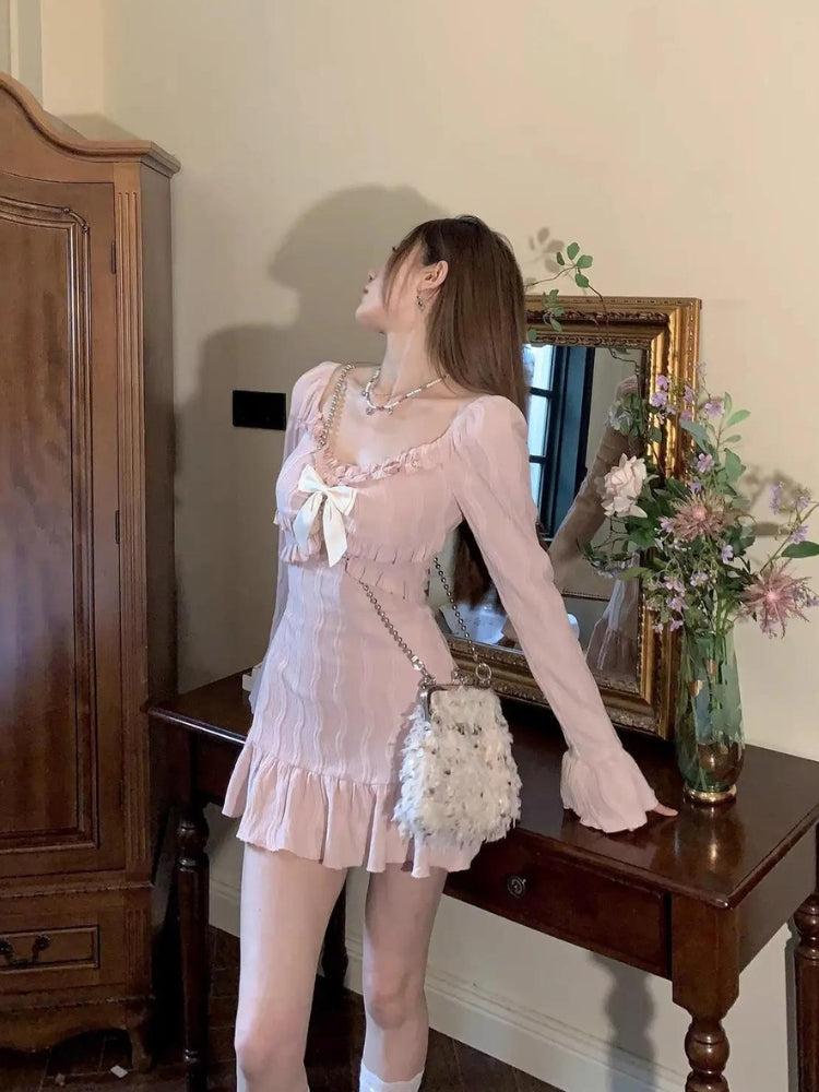Woman wearing a pink dress with a bow and ruffles, standing next to a table with flowers.