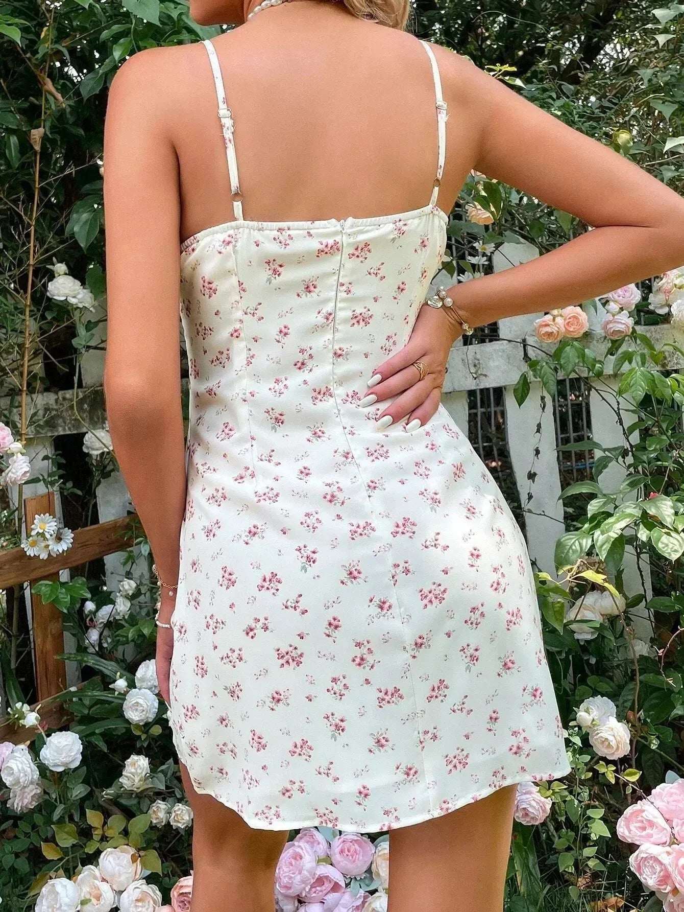 Back view of a woman wearing a white floral print mini dress with spaghetti straps and a tie front, in a garden.