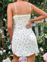 Back view of a woman wearing a white floral print mini dress with spaghetti straps and a tie front, in a garden.