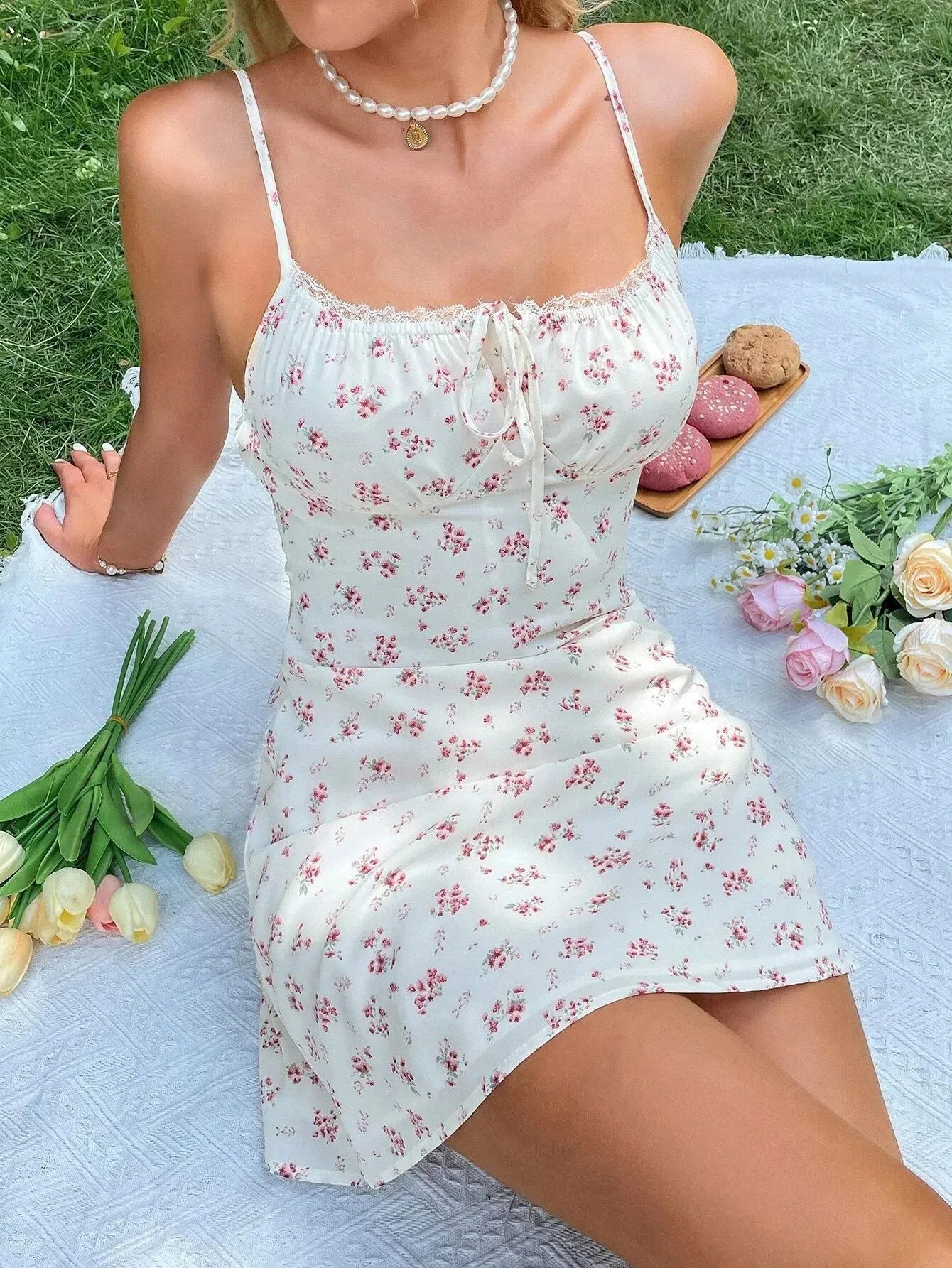 Woman wearing a white floral print mini dress with spaghetti straps and a tie front, sitting on a picnic blanket in the grass.