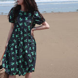 A woman poses on a beach wearing a black dress with a green floral print and puff sleeves.